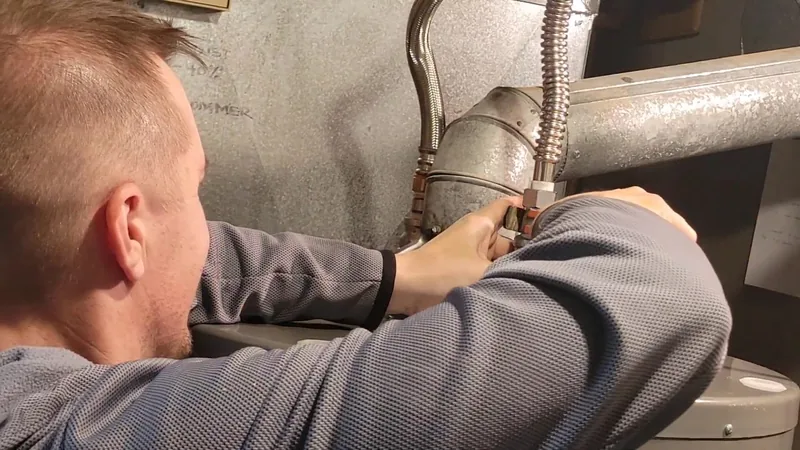 A man in a gray sweater reaching over the top of a tank water heater to work on its supply line